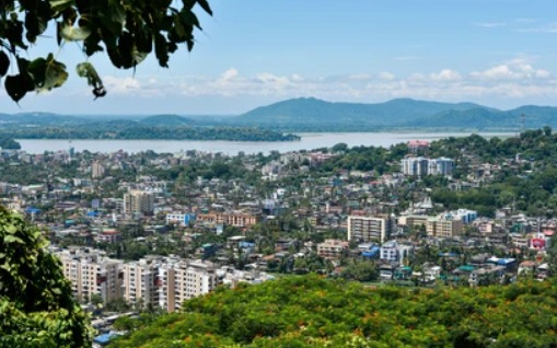 “Aerial view of Guwahati city with buildings, green hills, and the Brahmaputra river under a clear sky.”