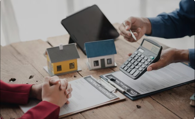 Bank official discussing property loan and mortgage calculations with a client, showing house models, calculator, and contract agreement on a wooden table